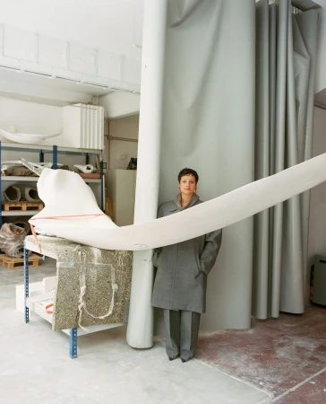 Woman in grey coat stands in an art studio with a large white abstract sculpture and white curtain. Indoor setting, neutral color palette.