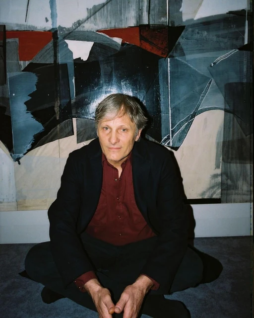 Elderly man in a red shirt and black jacket poses seated against an abstract geometric art background in a photo studio setting.