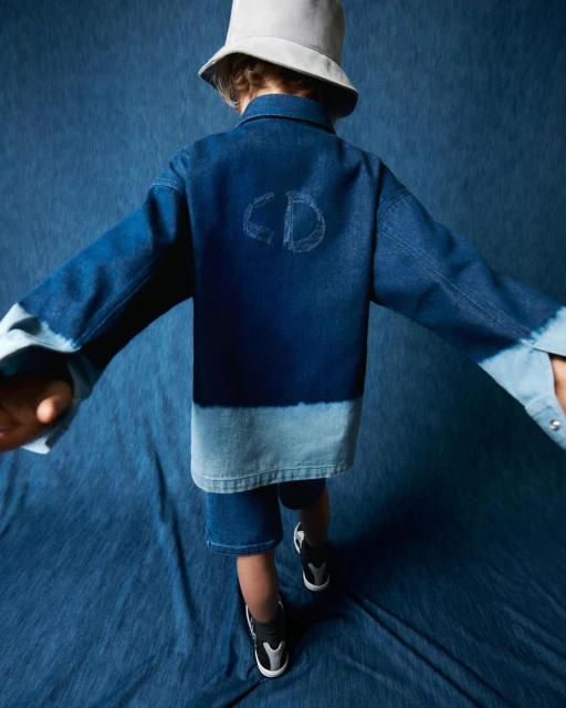 Child in denim jacket with CD logo and matching shorts stands against a blue textured background, wearing a grey bucket hat and sneakers.