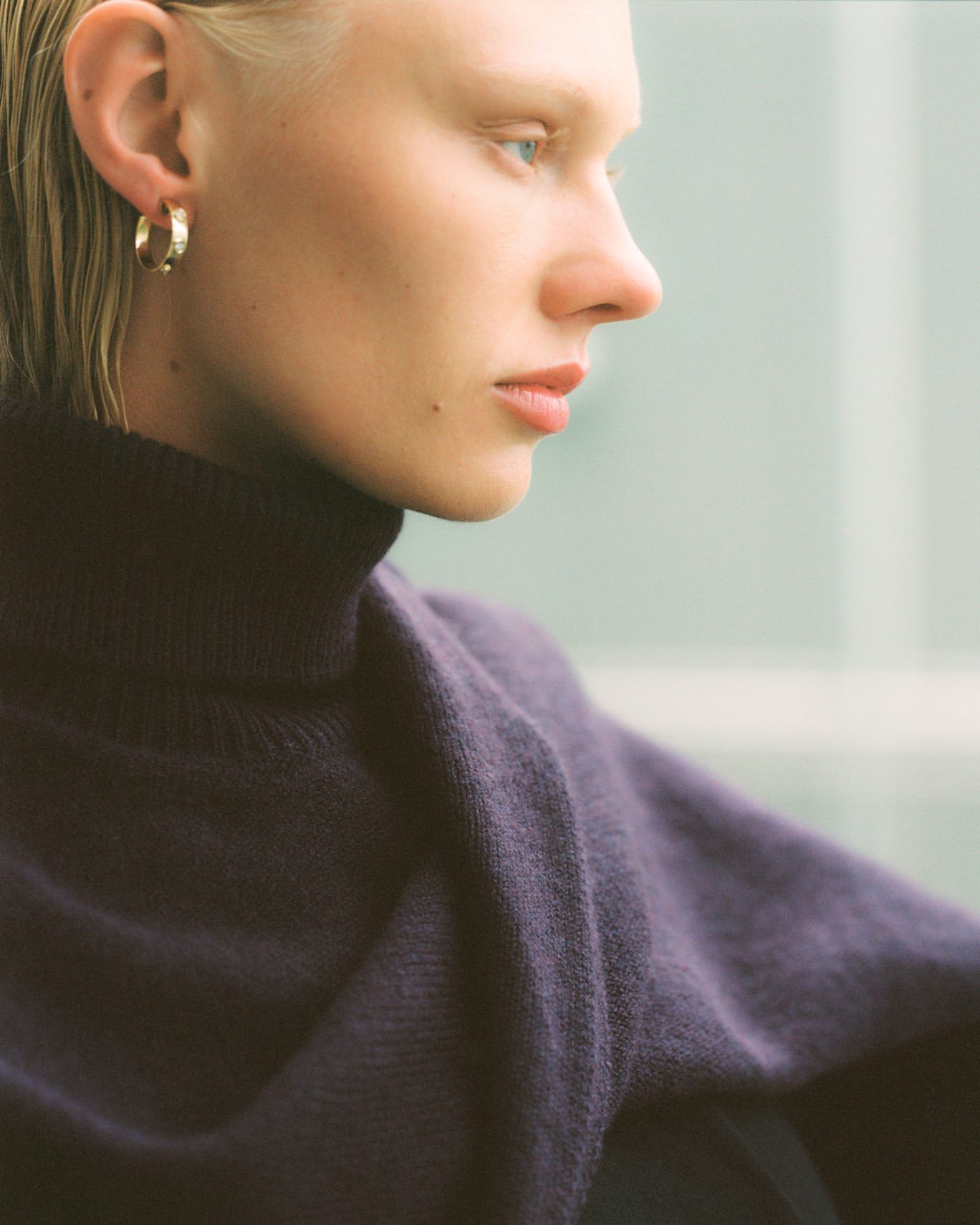 Close-up portrait of a light-skinned adult female with short blonde hair, wearing a purple sweater and gold earrings, against a blurred background.