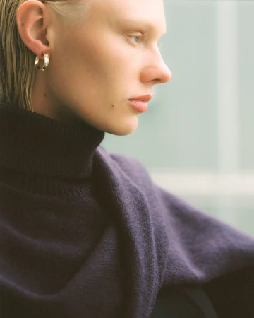 Close-up portrait of a light-skinned adult female with short blonde hair, wearing a purple sweater and gold earrings, against a blurred background.