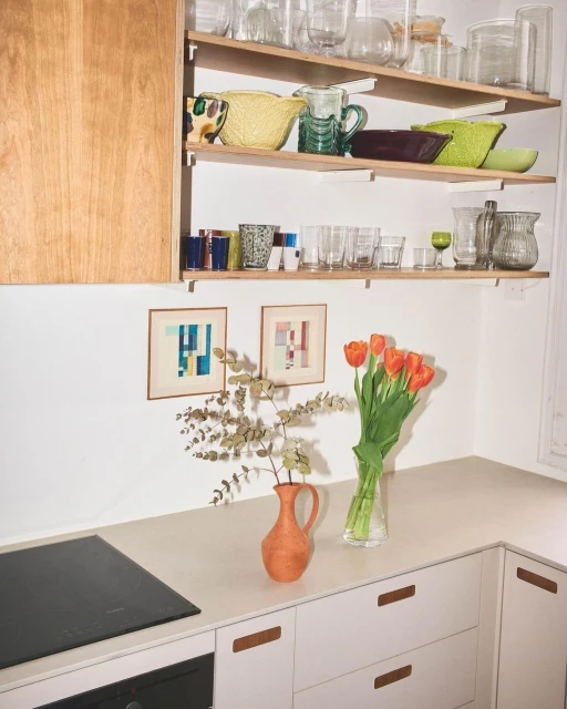 Bright, modern kitchen interior with open shelving displaying glassware, orange tulips in a vase, and abstract artwork on the wall.
