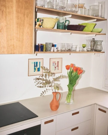 Bright, modern kitchen interior with open shelving displaying glassware, orange tulips in a vase, and abstract artwork on the wall.