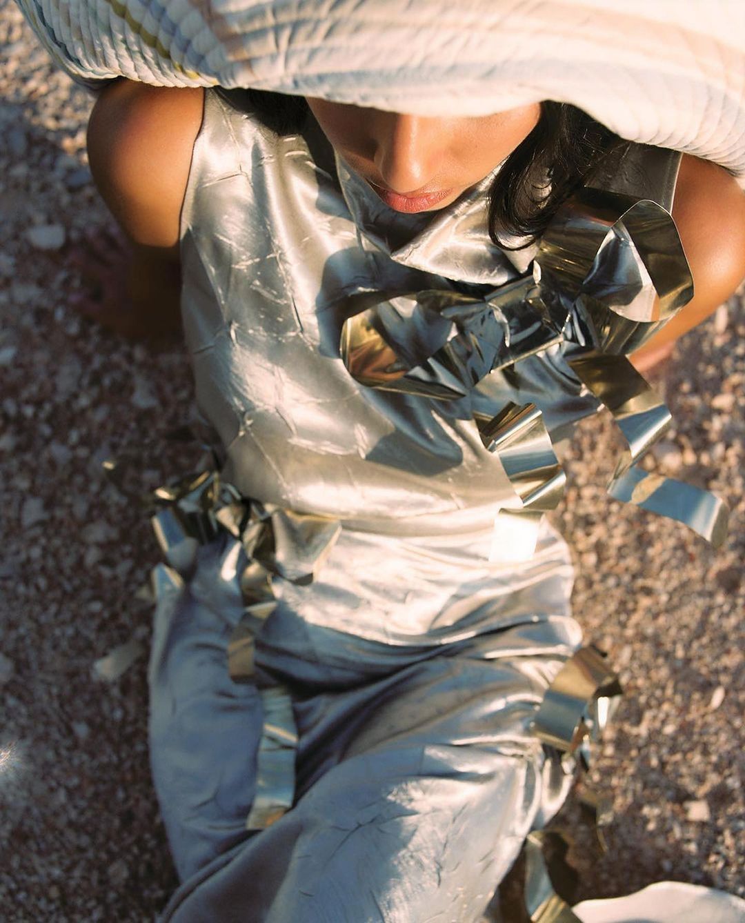 A woman wearing a silver satin outfit and a large hat, posing in an outdoor desert setting with scattered silver confetti. 