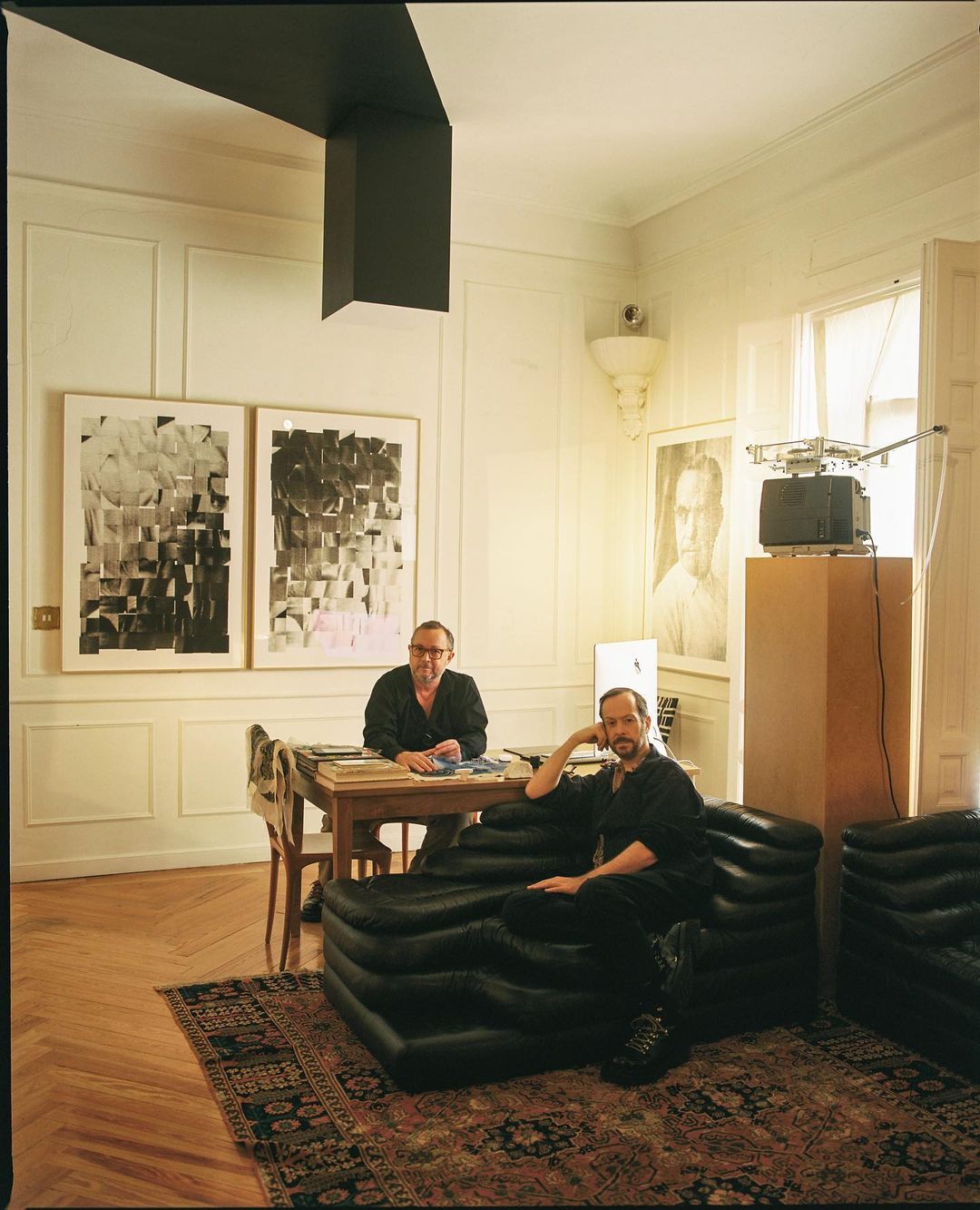 Indoor portrait of two adult males seated in a room with modern art, black furniture, and a vintage television set on a pedestal.