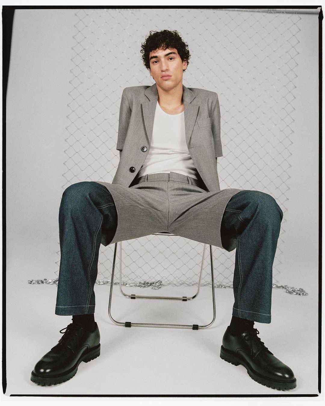 Fashion shot of a young adult male model sitting on a chair, wearing a grey suit jacket, white t-shirt, denim shorts, black socks, and black shoes.