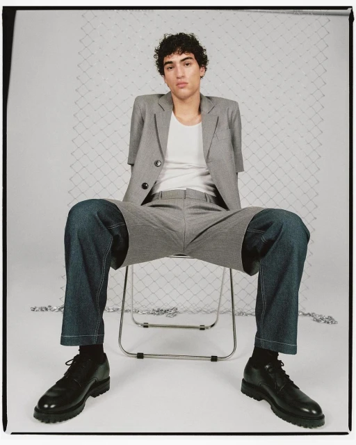 Fashion shot of a young adult male model sitting on a chair, wearing a grey suit jacket, white t-shirt, denim shorts, black socks, and black shoes.