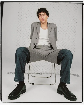 Fashion shot of a young adult male model sitting on a chair, wearing a grey suit jacket, white t-shirt, denim shorts, black socks, and black shoes.