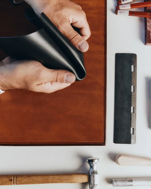 Hands holding black leather in a crafting scene with tools, a brown leather surface, and a white background on a flat lay.