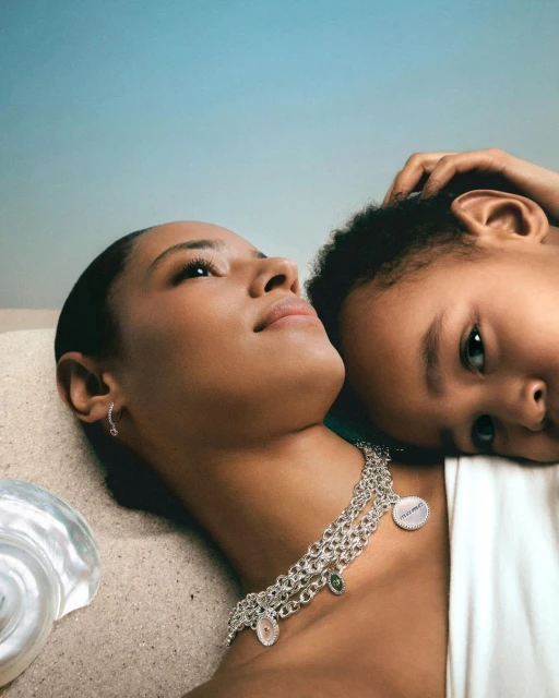 Elegant portrait of an adult female and a child lying on a sandy surface, both wearing stylish silver necklaces with dangling charms.