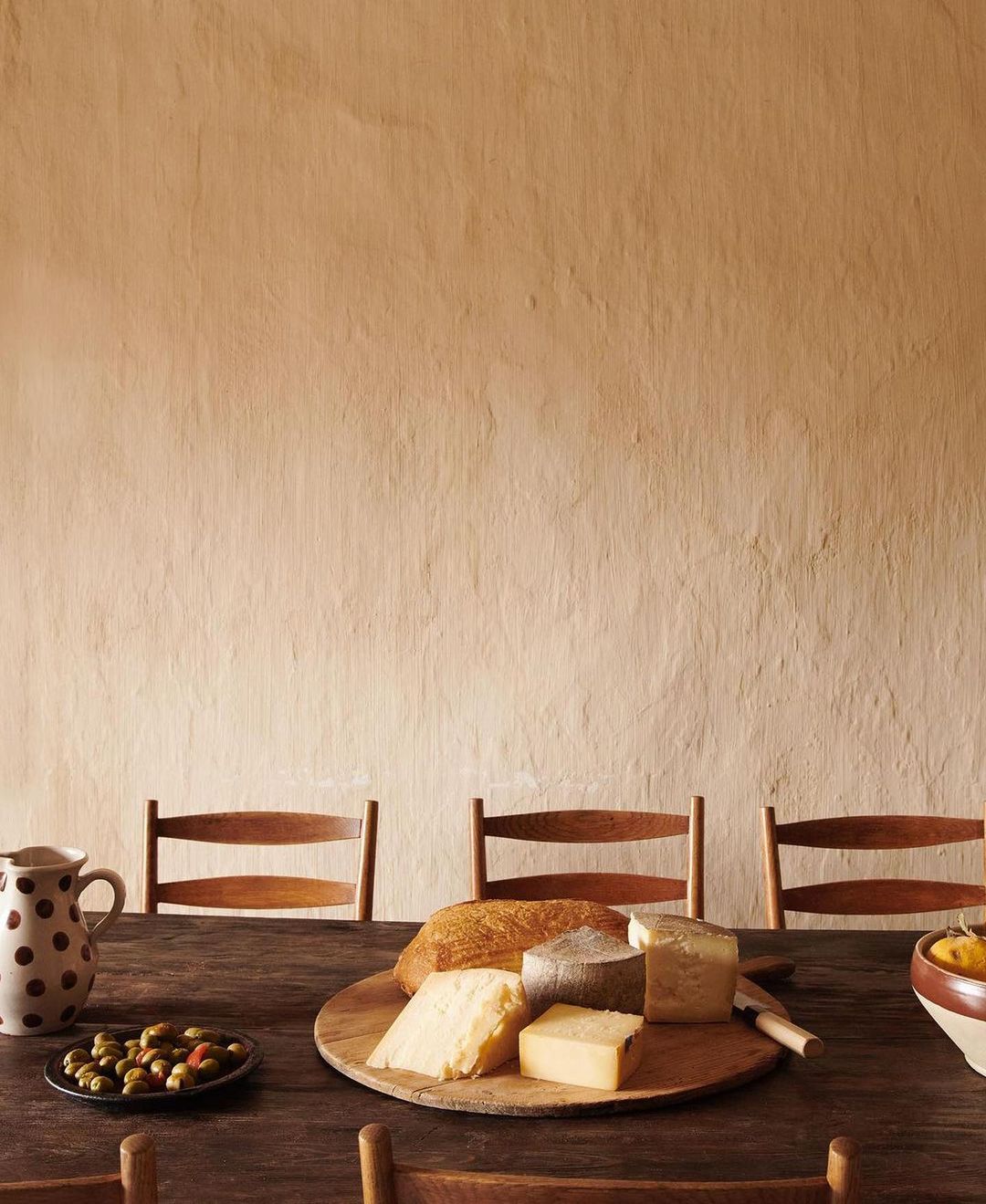 Still life shot of a wooden table setting with cheese, bread, olives, a pitcher, and chairs against a textured neutral wall.