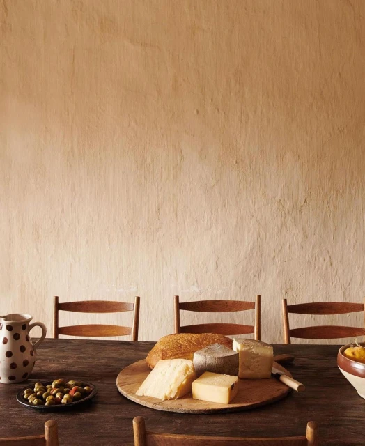 Still life shot of a wooden table setting with cheese, bread, olives, a pitcher, and chairs against a textured neutral wall.