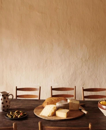 Still life shot of a wooden table setting with cheese, bread, olives, a pitcher, and chairs against a textured neutral wall.