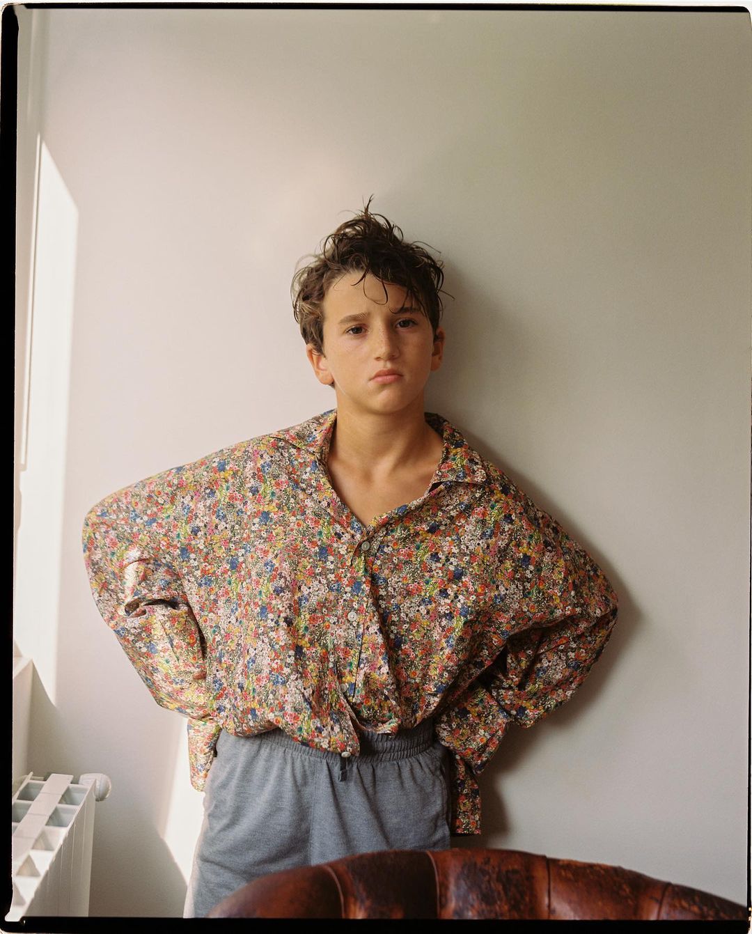 Teen boy in a floral shirt and grey shorts stands indoors against a white wall with natural lighting and an old leather chair.