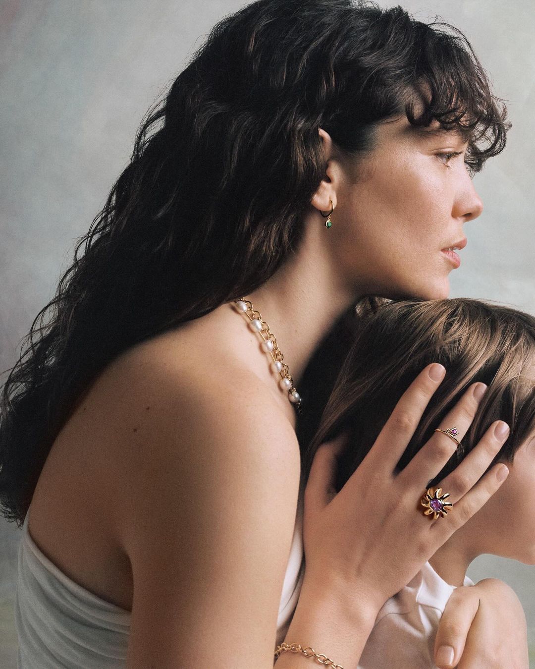 Portrait of a woman holding a child in profile, wearing gold jewelry, a pearl necklace, and a white top against a textured wall.