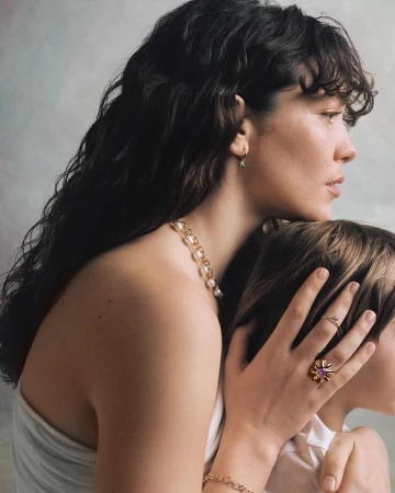Portrait of a woman holding a child in profile, wearing gold jewelry, a pearl necklace, and a white top against a textured wall.