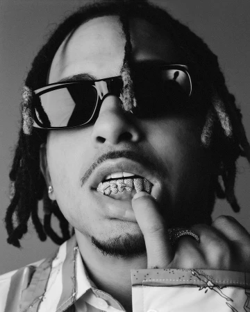 Black and white portrait of adult male wearing sunglasses, patterned shirt, grill, and dreadlocks, with finger touching teeth.