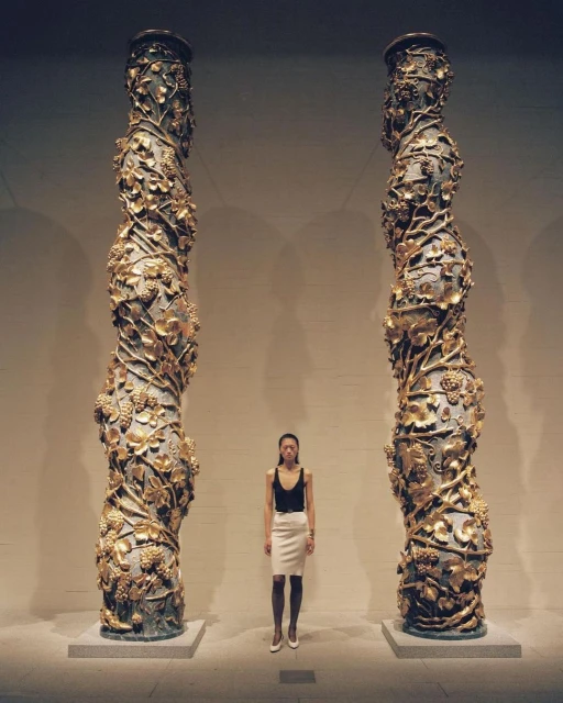 Elegant woman stands centered between two large, gold-accented, vine-covered art columns indoors against a neutral wall, showcasing art and fashion.