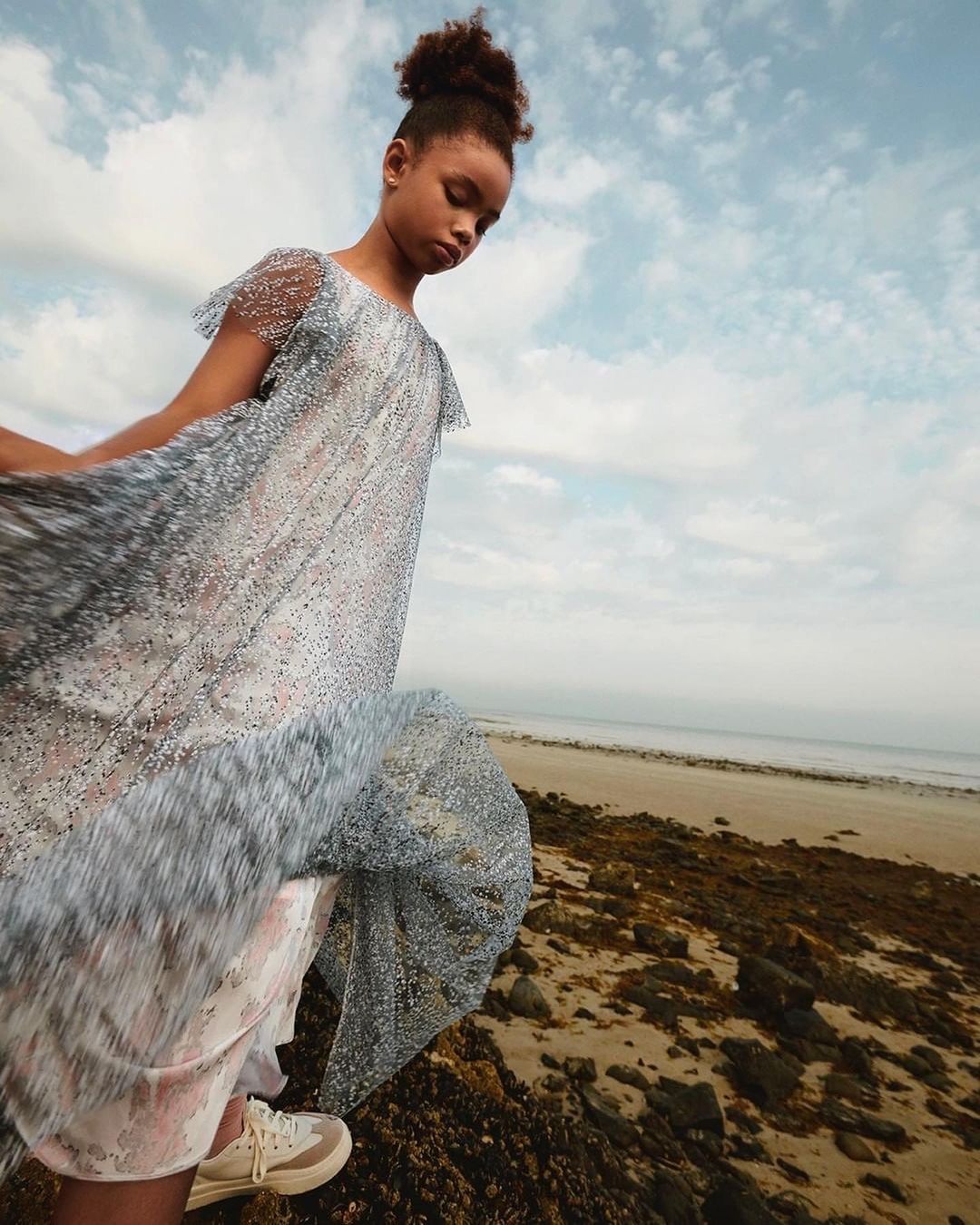 Young girl in a shimmering silver dress standing on a rocky beach under a cloudy sky, photographed by Pablo Zamora.