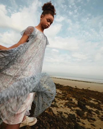 Young girl in a shimmering silver dress standing on a rocky beach under a cloudy sky, photographed by Pablo Zamora.