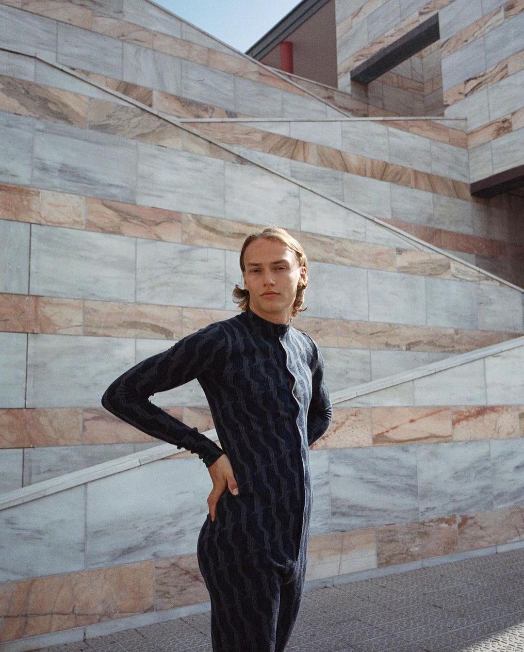 Young adult poses in dark patterned jumpsuit against a textured marble wall with steps, creating an editorial outdoor fashion portrait.