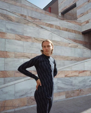 Young adult poses in dark patterned jumpsuit against a textured marble wall with steps, creating an editorial outdoor fashion portrait.