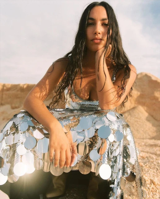 Attractive woman with long dark hair poses in a shimmering silver sequin dress in a desert landscape, hand on cheek, medium shot.