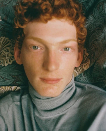 Close-up portrait of a young man with curly red hair and green eyes wearing a grey turtleneck against a patterned background.