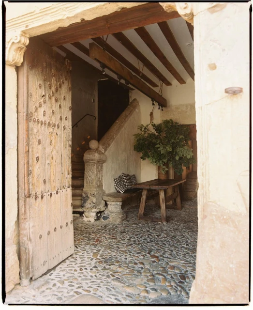 Rustic stone entryway features an open wood door, cobblestone floor, wooden beams, and a bench with greenery near a staircase.
