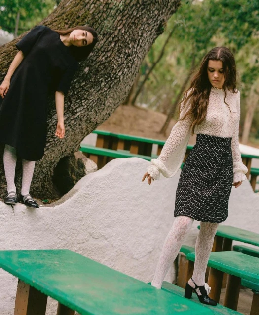 Two women in dresses posing outdoors near a tree and green picnic tables, showcasing fashion and style in a natural setting.