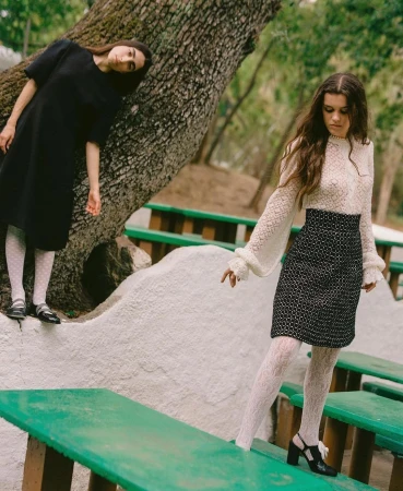 Two women in dresses posing outdoors near a tree and green picnic tables, showcasing fashion and style in a natural setting.