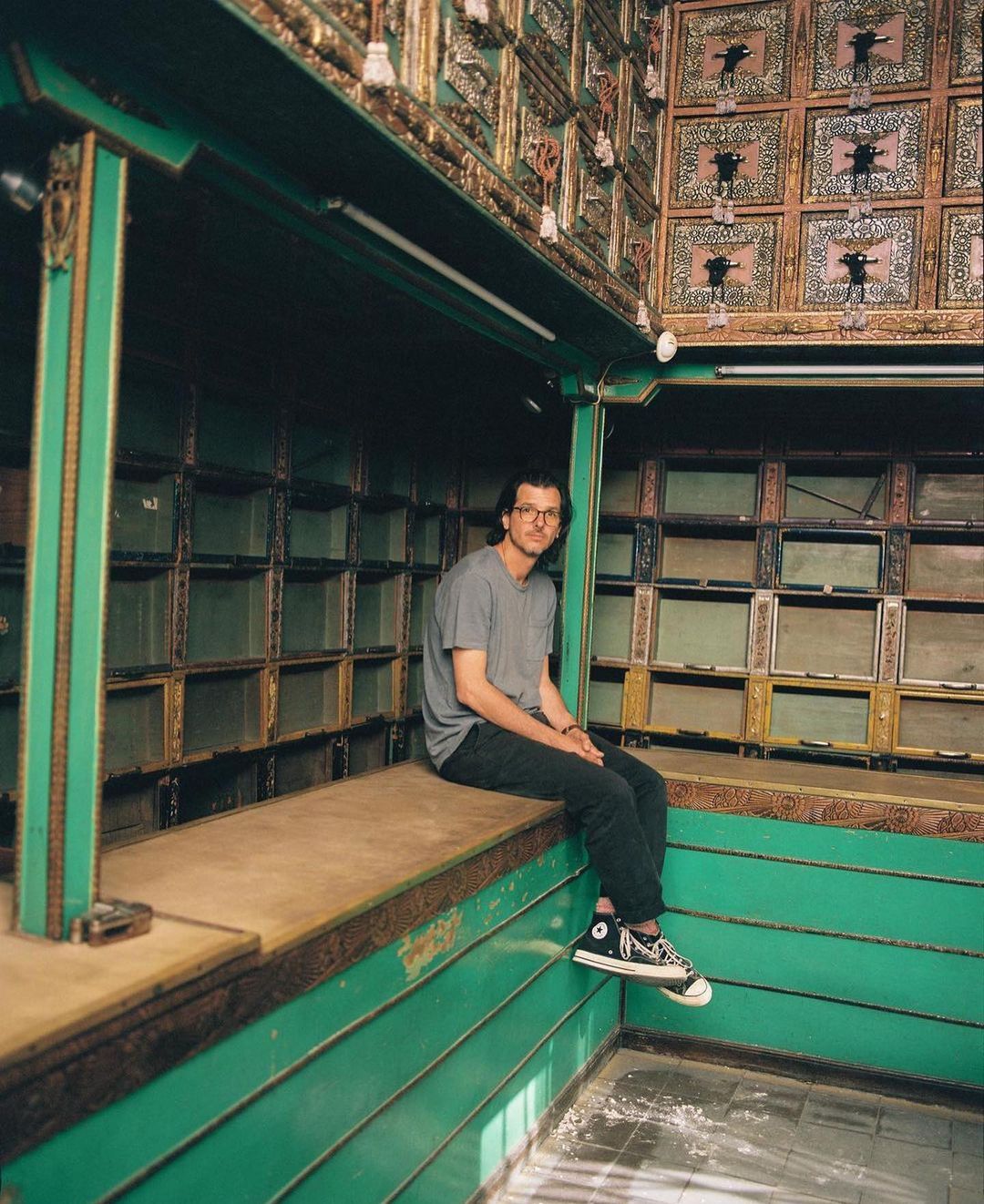 Young man sits on a green ledge inside an ornately decorated building with paneled walls and ceiling details in a medium shot.