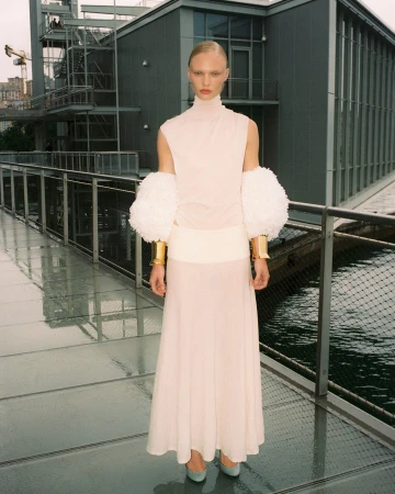 Fashion shoot: A woman in a light pink dress and large white arm cuffs stands on a glass walkway with a modern building in the background.