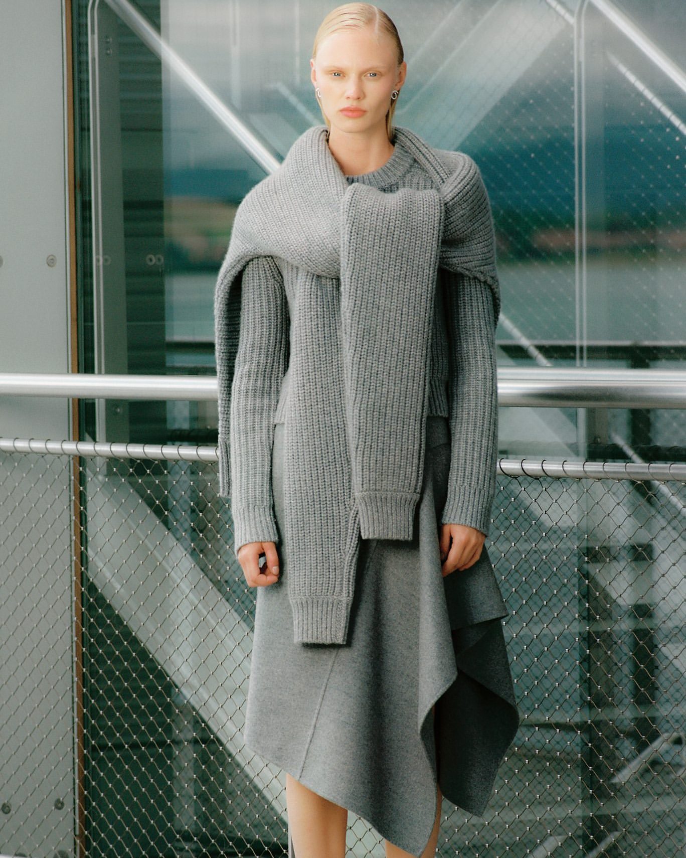 Woman in a gray knitted sweater and skirt stands in front of a glass wall. The neutral color palette emphasizes texture and minimalist style.