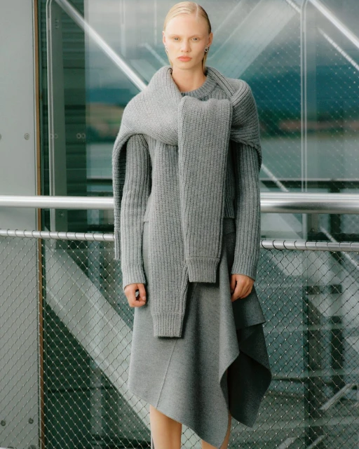 Woman in a gray knitted sweater and skirt stands in front of a glass wall. The neutral color palette emphasizes texture and minimalist style.