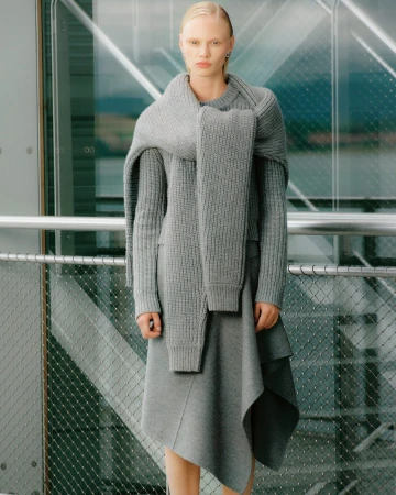 Woman in a gray knitted sweater and skirt stands in front of a glass wall. The neutral color palette emphasizes texture and minimalist style.