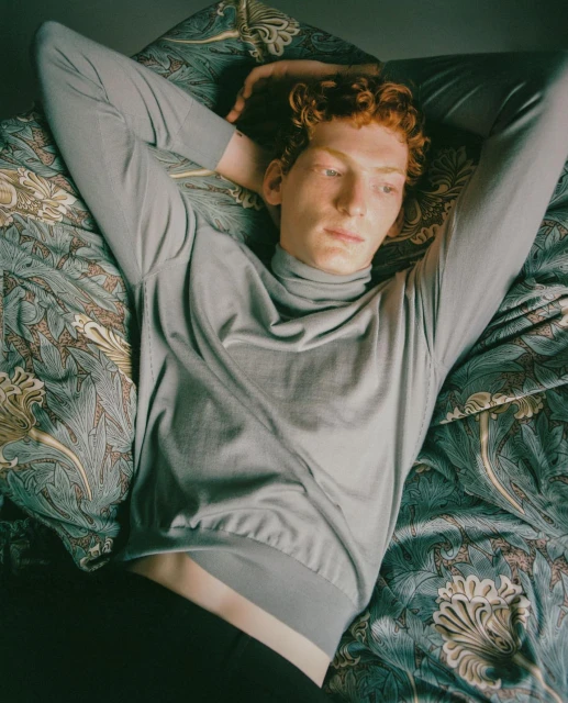 Red-haired man in gray turtleneck lying on a patterned bedspread, eyes closed, arms above his head, looking serene.