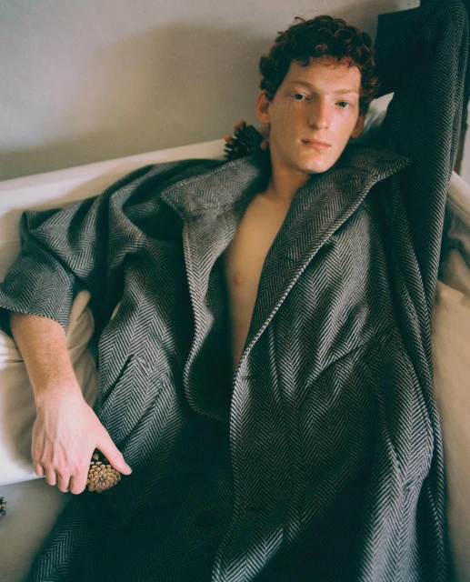 A young adult with red curly hair is lying down wearing a gray herringbone patterned coat, indoors. The lighting is soft and natural.