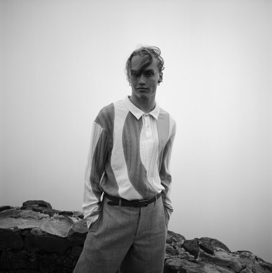 Young man in a striped polo shirt and trousers posing against a hazy outdoor background with rocks