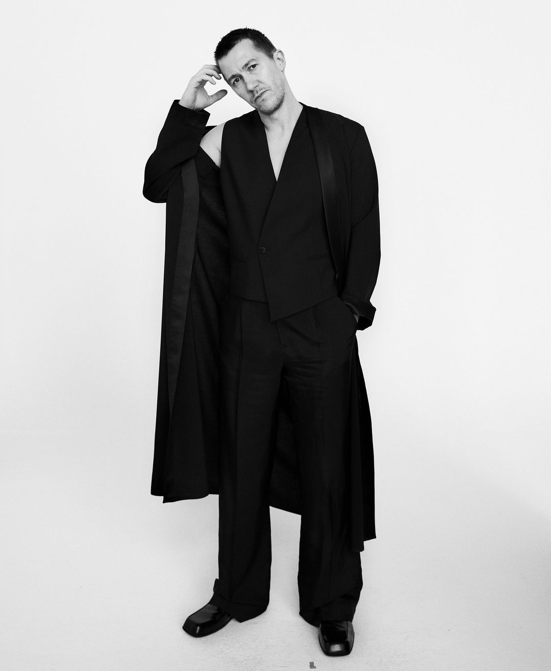 Man wearing a black suit and long coat with one sleeve slipped off, posing against a white background.
