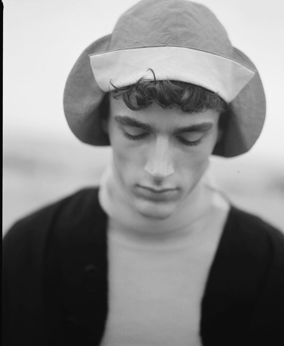 Monochrome portrait of a young adult male wearing a vintage-style hat and turtleneck, looking down with a neutral expression in an outdoor setting.
