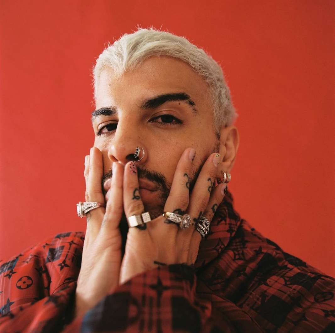 Portrait of a stylish adult male with bleached hair, facial piercings, tattoos, and rings posing in front of a bright red background.