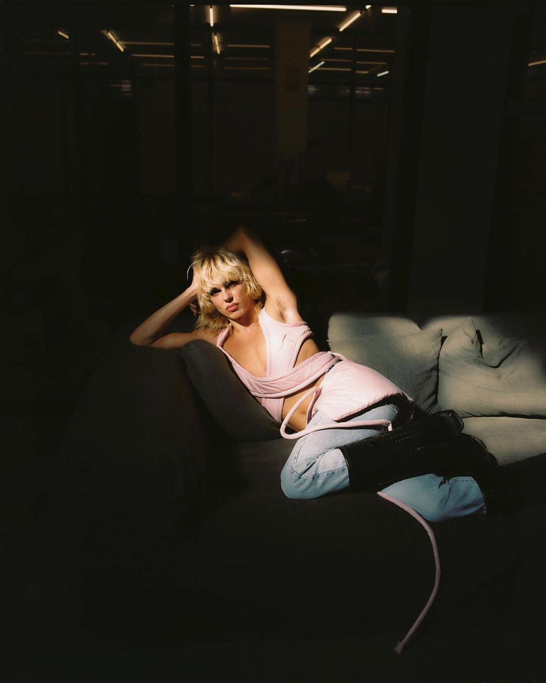 Adult female laying on grey couch in a pink top and light wash jeans in a dark room with artificial lighting overhead.