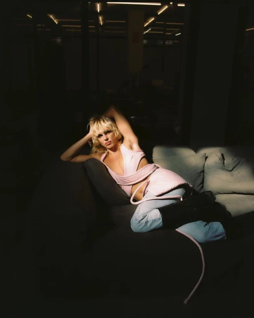 Adult female laying on grey couch in a pink top and light wash jeans in a dark room with artificial lighting overhead.