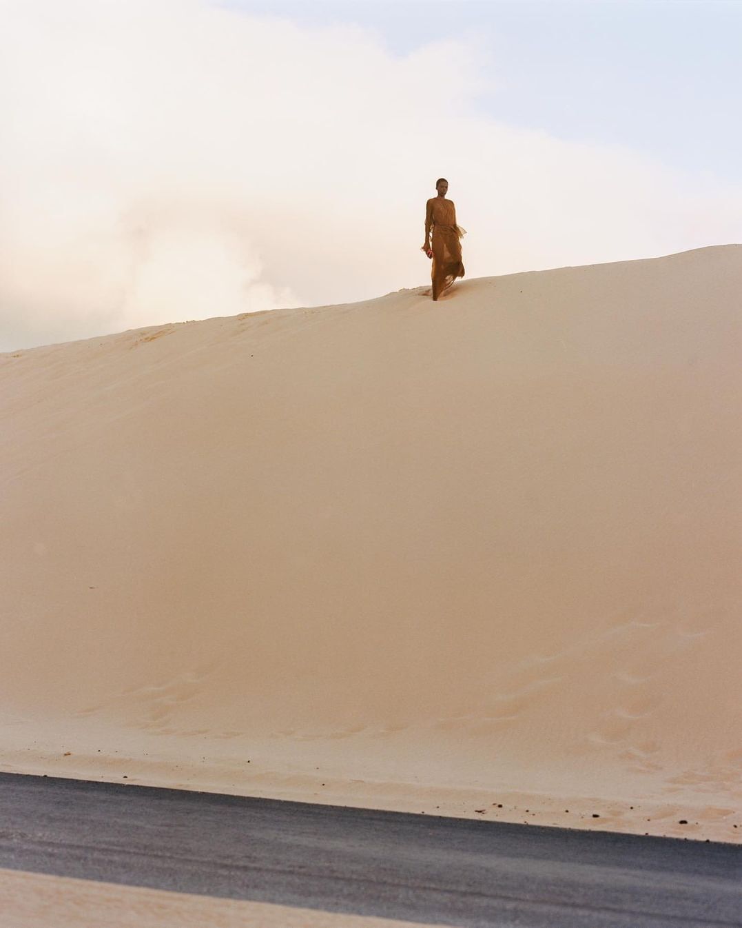 Figure standing atop a large sandy dune above a paved road under an overcast sky, captured in a wide landscape shot.