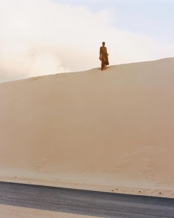 Figure standing atop a large sandy dune above a paved road under an overcast sky, captured in a wide landscape shot.