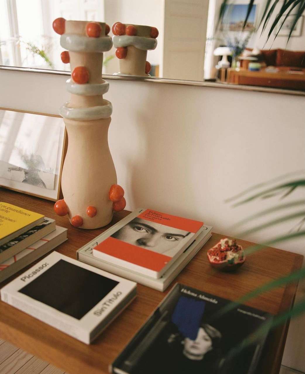 Still life of ceramic vases with orange spheres, stacked books, and a pomegranate on a wooden table, indoor setting.