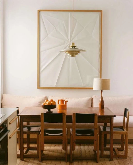 Indoor dining area features wooden table and chairs, bowl of oranges, orange pitcher, lamp, and geometric wall art with pendant light.