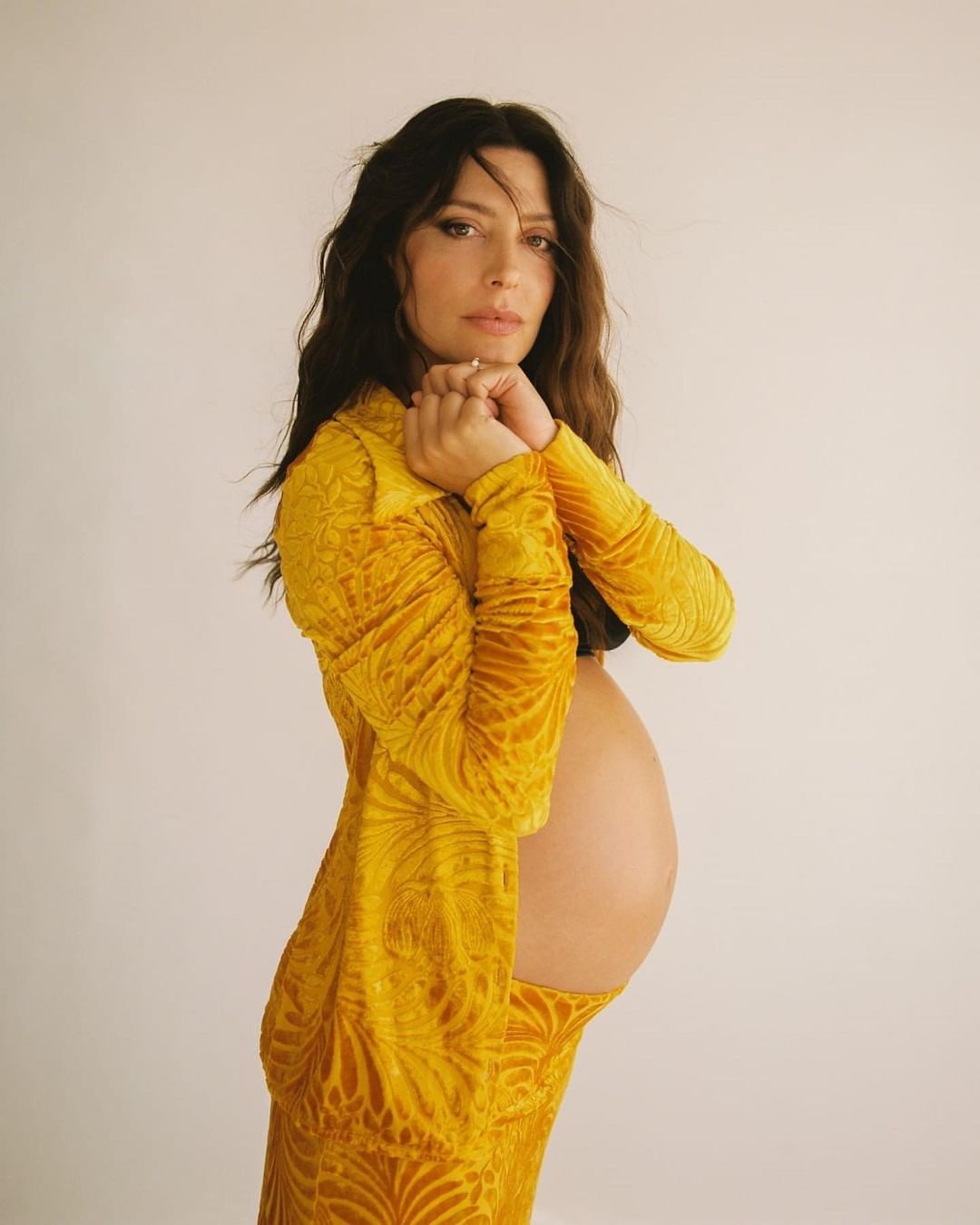 Portrait of a pregnant adult woman in a textured yellow dress. She has long dark hair and her hands are clasped under her chin.