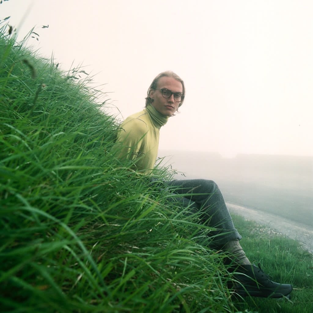 Man with glasses sits on a grassy hill in a yellow turtleneck and gray jeans, looking at the camera with fog in the distance.